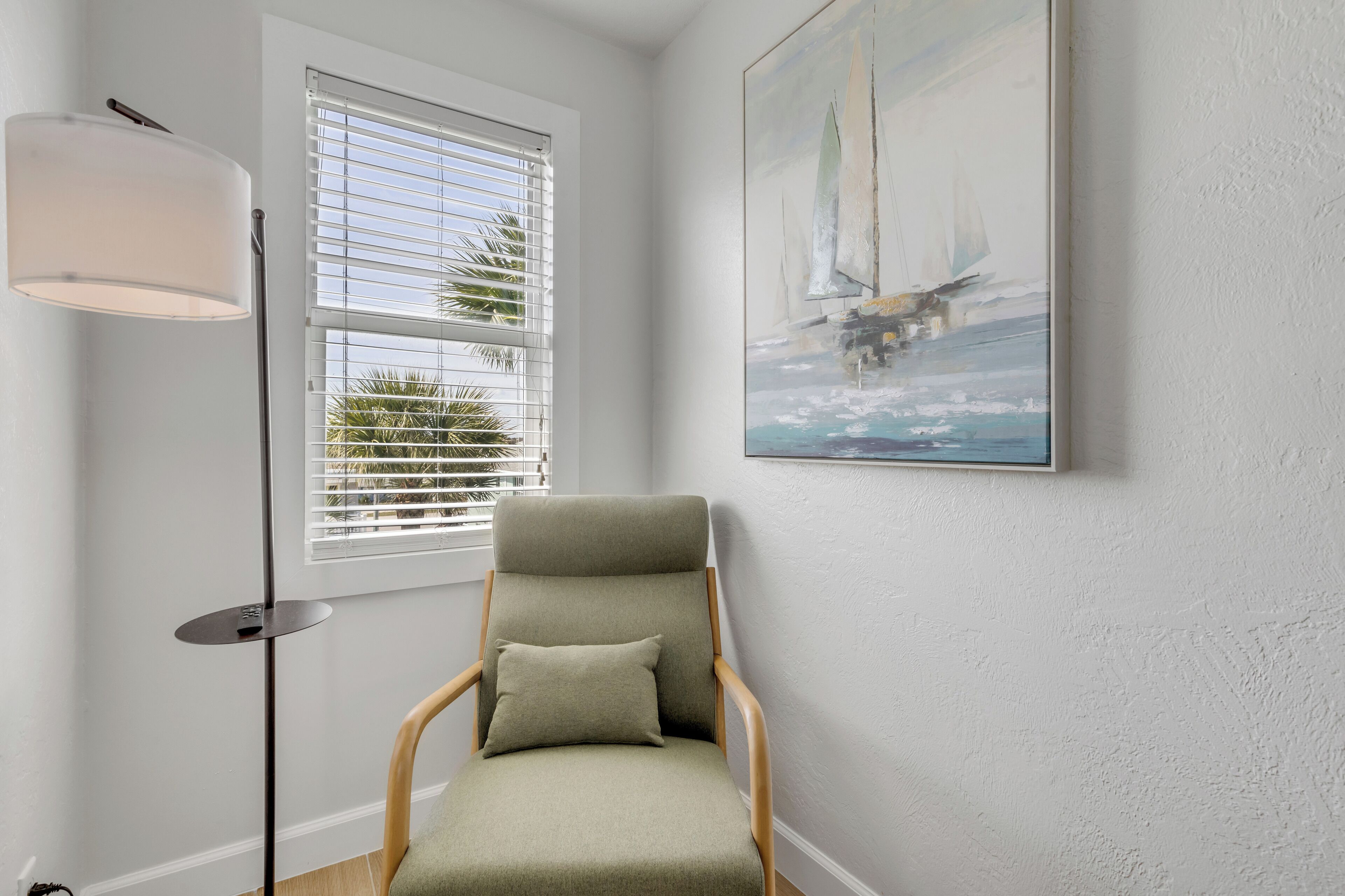 A room with a chair, a floor lamp, a window with blinds, and a painting of a sailboat.