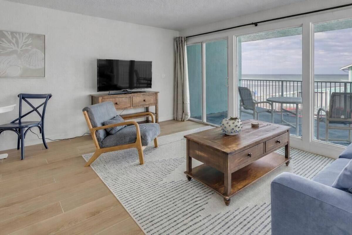Uma sala de estar moderna com uma grande porta de vidro deslizante que dá para uma varanda com vista para a praia e uma casa.