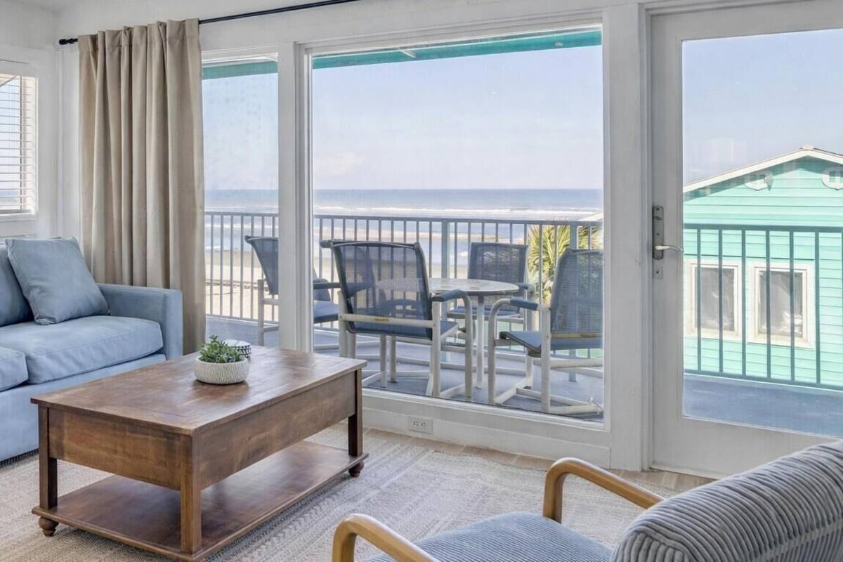 Uma sala de estar com um sofá, uma mesa de centro e vista para a praia através de uma porta de vidro deslizante.