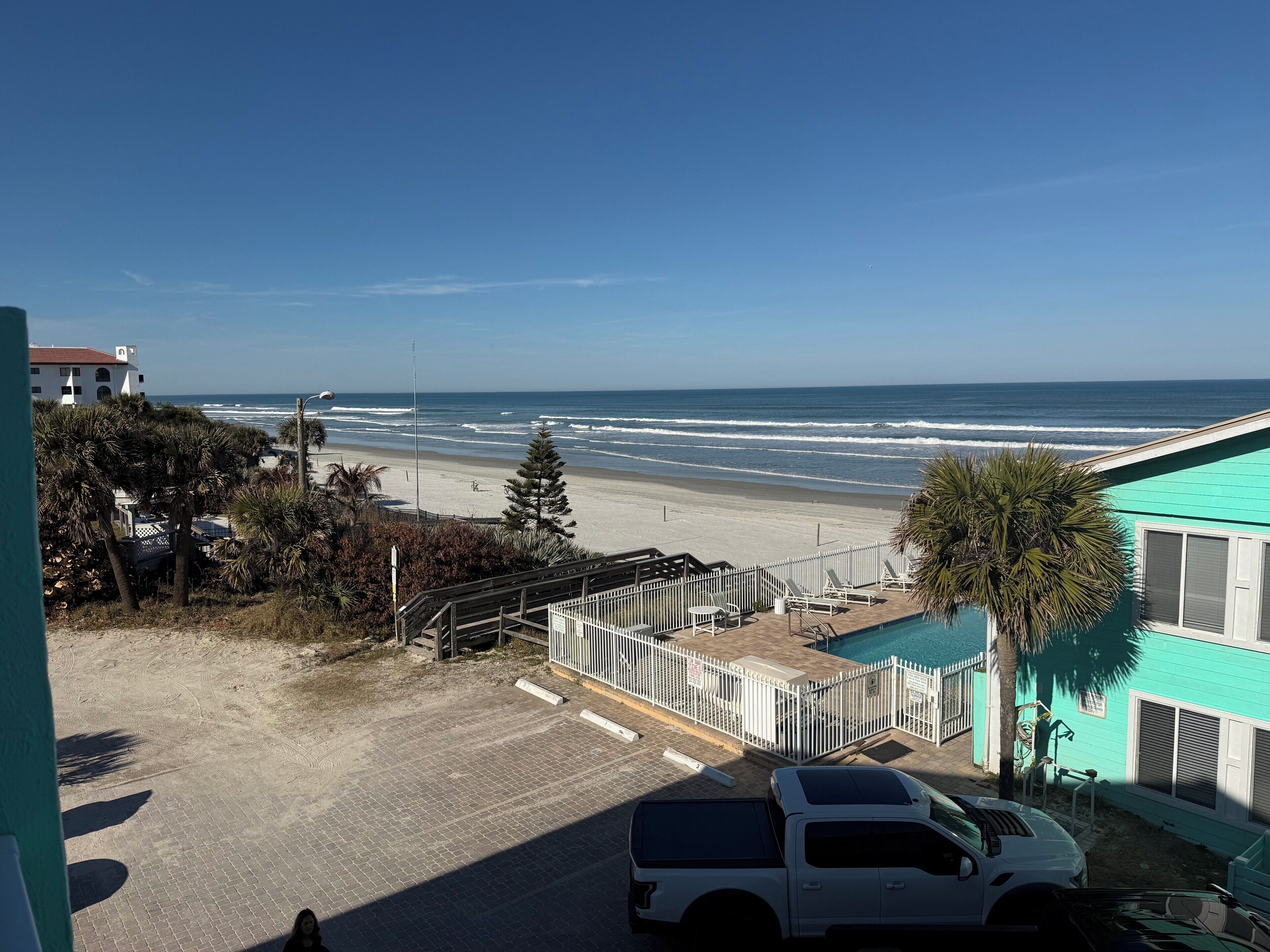 Vista de una playa con un área de estacionamiento, una piscina y un edificio de fachada turquesa.