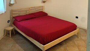 A single bed with a wooden frame and red bedding in a room with white walls and a tiled floor.