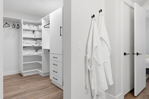 Ein moderner, weißer Raum mit Holzboden, einer eingebauten Garderobe mit mehreren Regalen und Schubladen sowie einem auf einem Haken aufgehängten Bademantel.