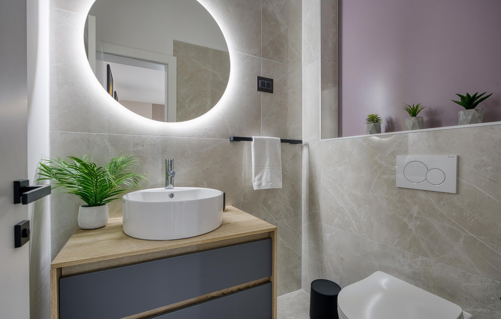 Une salle de bain moderne avec un miroir rond, un lavabo blanc, un porte-serviettes et des toilettes. On y trouve des plantes sur le comptoir et une petite poubelle.
