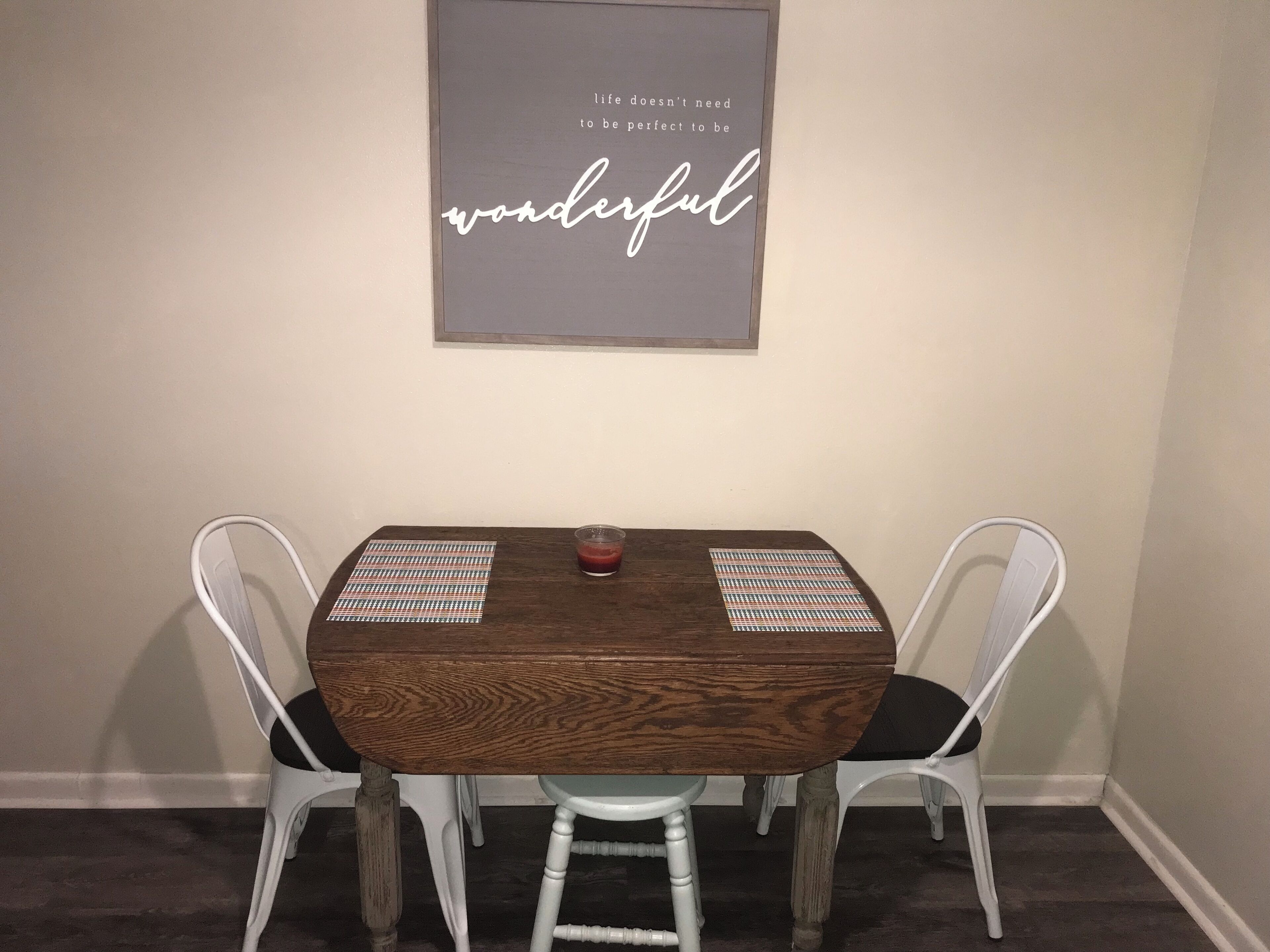 A dining table with two chairs, a framed quote on the wall, and a small glass on the table.
