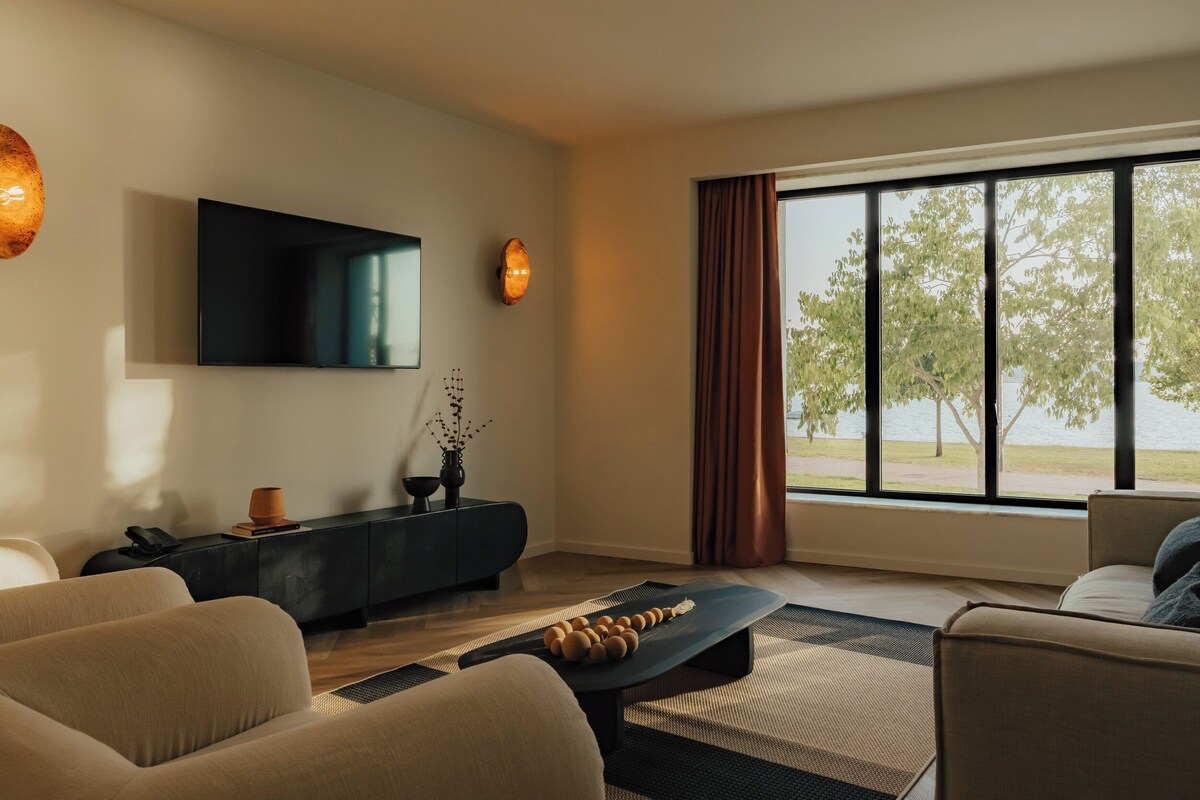Una sala de estar moderna con un televisor de pantalla plana, un mueble bajo oscuro, un sofá y un ventanal con vistas a árboles y agua.