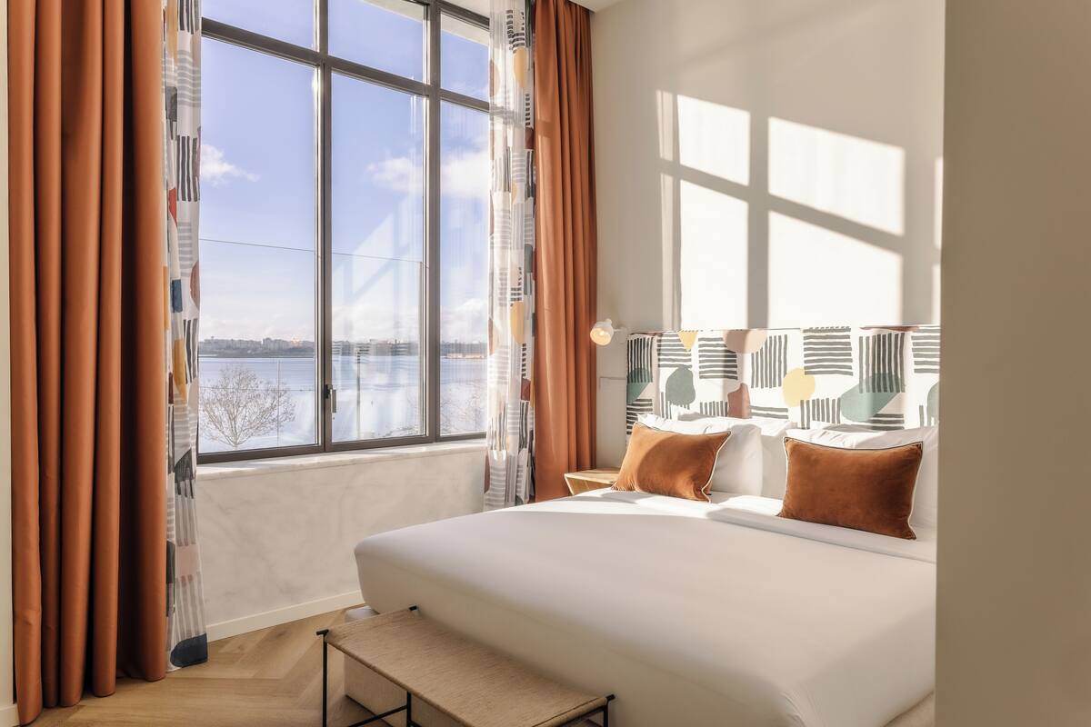 Een hotelkamer met een groot bed, uitzicht op de stad door een raam en een nachtkastje met een lamp.