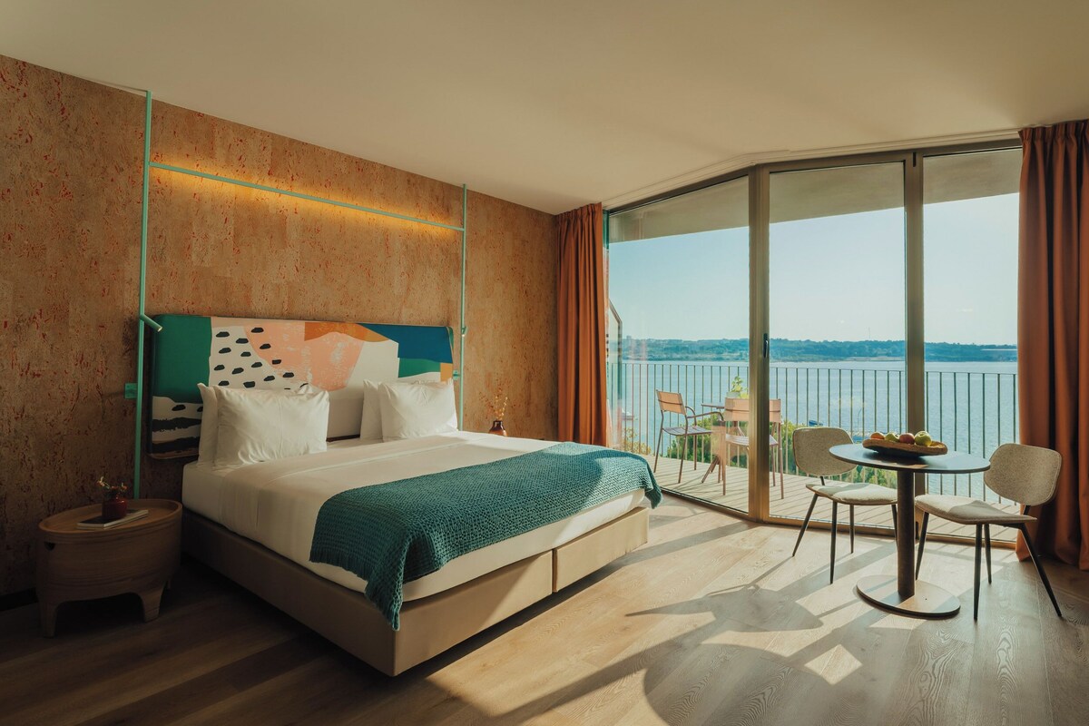 Habitación de hotel con cama, una pequeña mesa con sillas, un balcón con vistas y un ventanal grande.