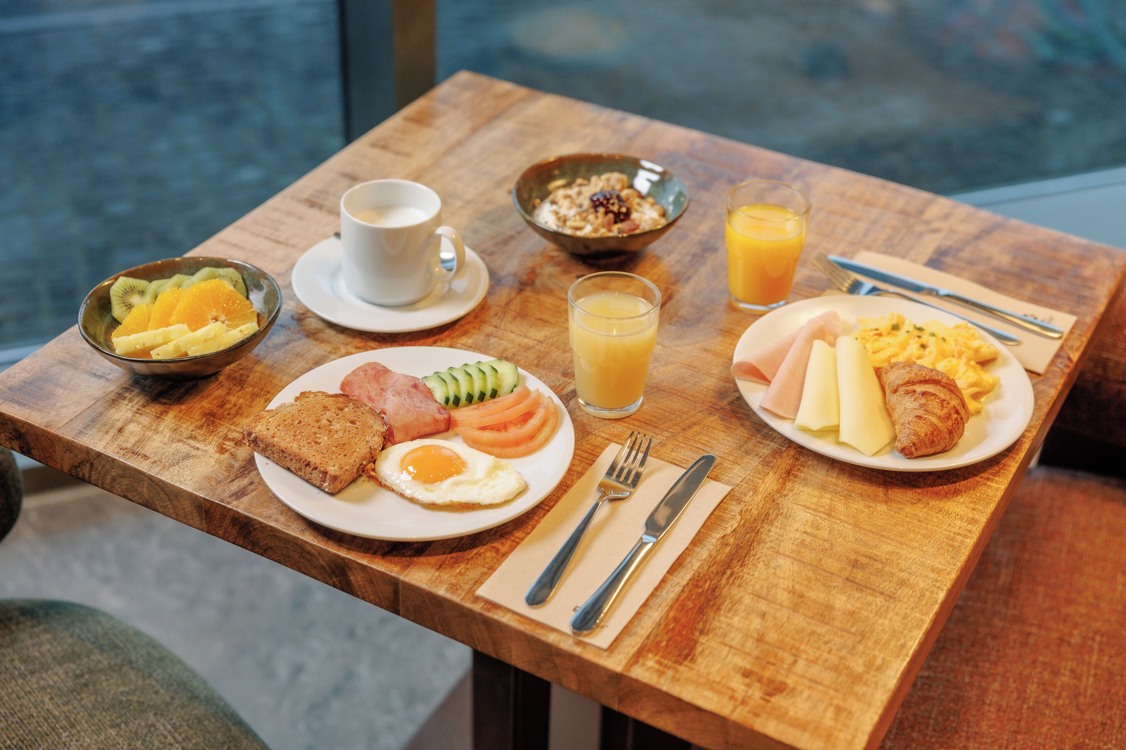 Una mesa de madera con productos para el desayuno, como tostadas, huevos, jamón y un croissant, además de un bol de fruta y un vaso de zumo de naranja.
