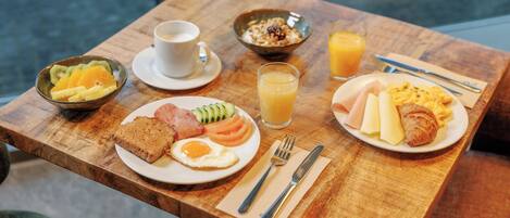 A wooden table with breakfast items including toast, eggs, ham, and a croissant, along with a bowl of fruit and a glass of orange juice.
