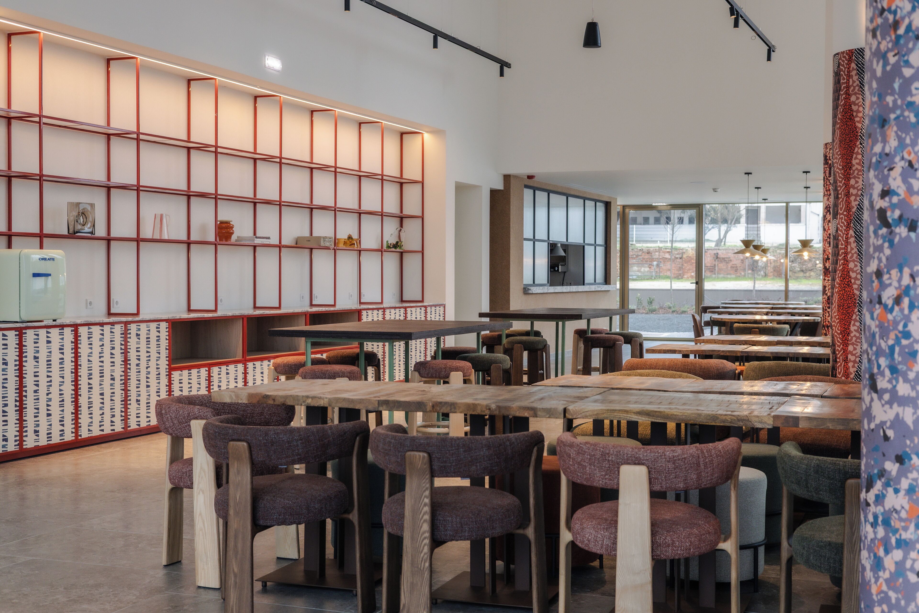 A modern restaurant interior with a long communal table, multiple chairs, and a wall-mounted shelf with decorative items.