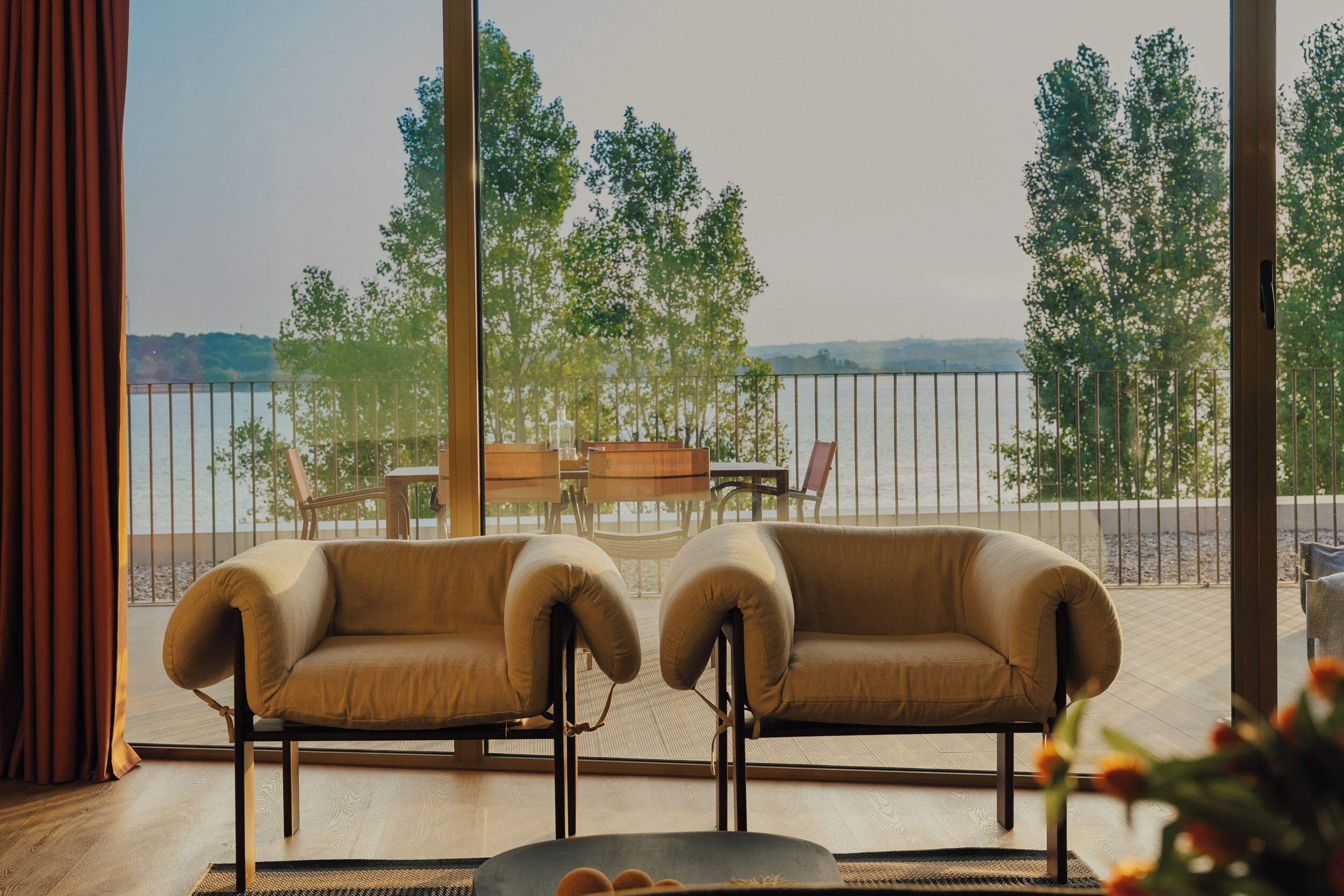 A room with a view of a body of water, two armchairs, a small table, and a view of trees.