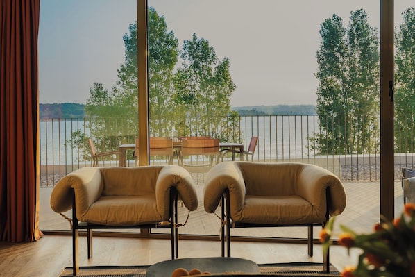 A room with a view of a body of water, two armchairs, a small table, and a view of trees.