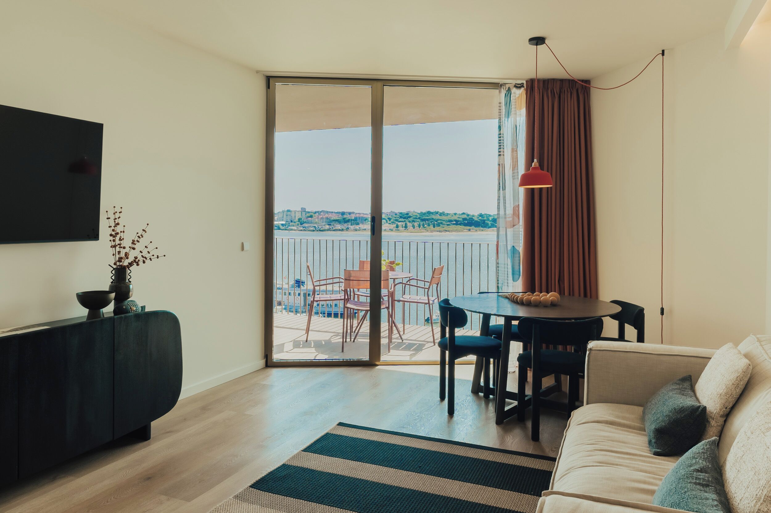 A modern living room with a sofa, a dining table with chairs, a TV mounted on the wall, and a view of a balcony with a table and chairs outside.