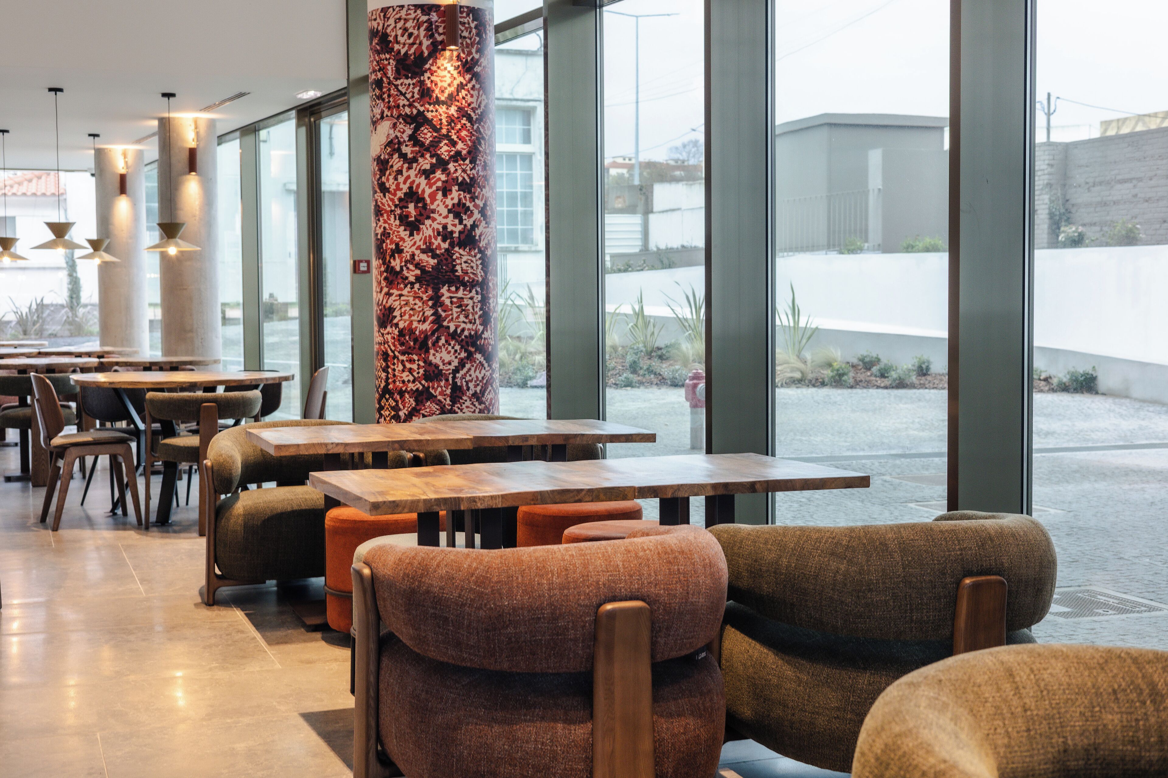 En modern restauranginteriör med träbord och stolar, stora fönster och en dekorativ pelare.