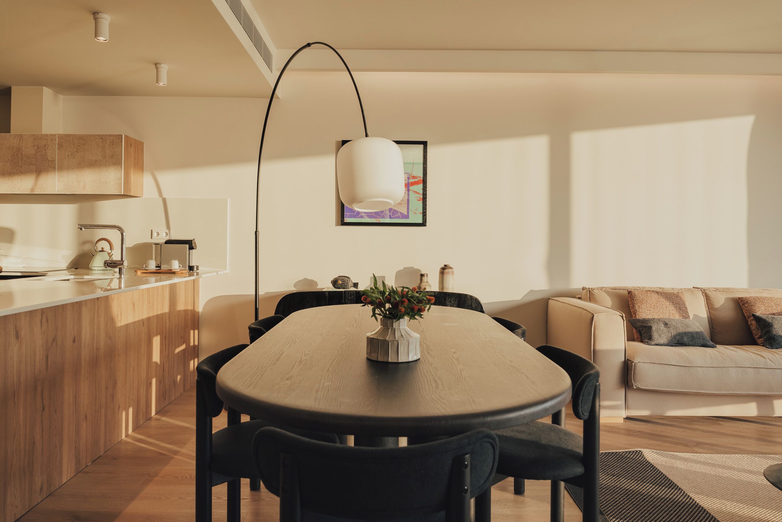 Uma sala de estar moderna com área de jantar, com uma mesa de jantar de madeira, cadeiras pretas, luminária pendente branca e um sofá bege com almofadas estampadas.