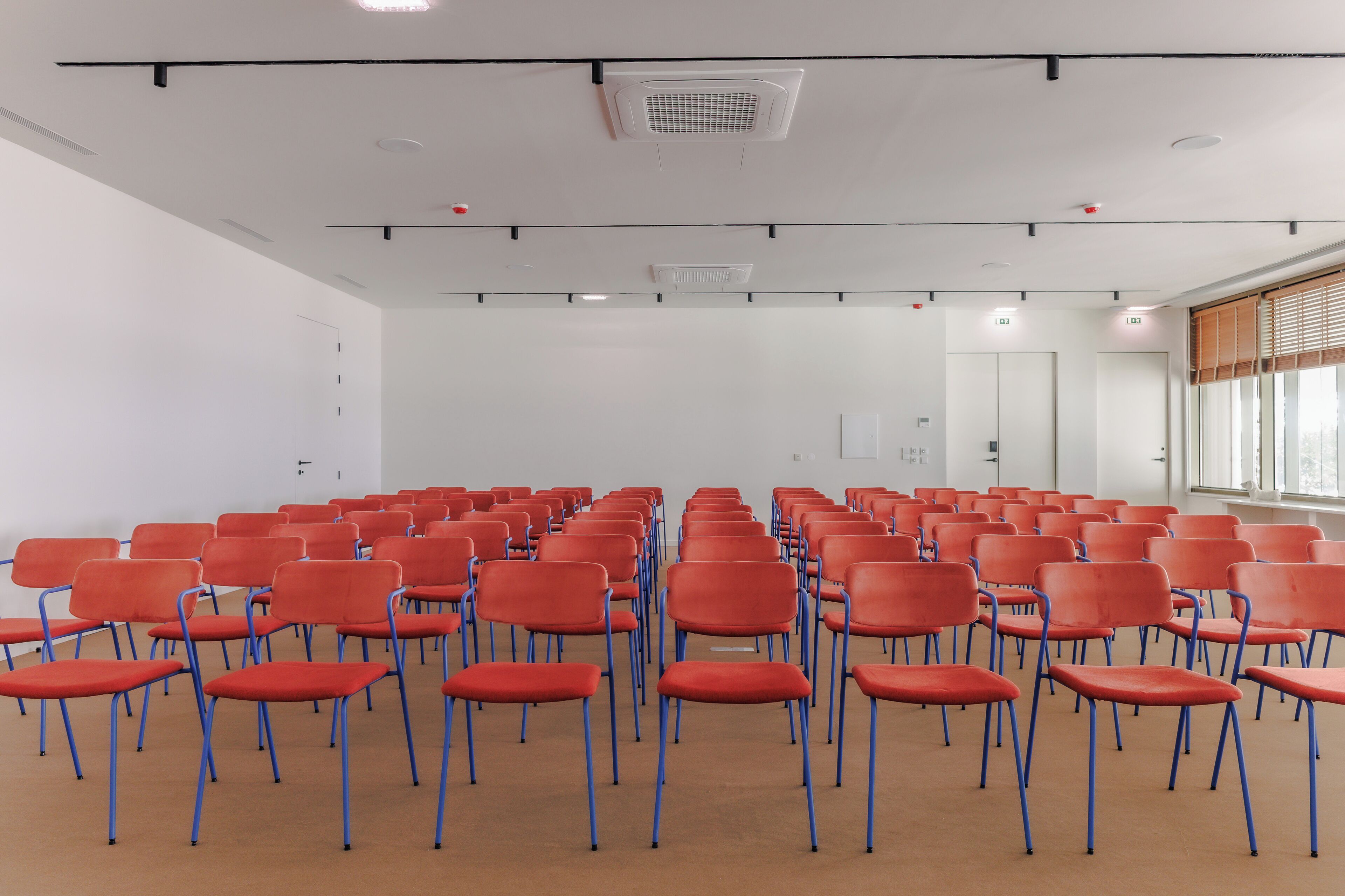 Un salón de conferencias con filas de sillas rojas y un amplio ventanal con persianas.