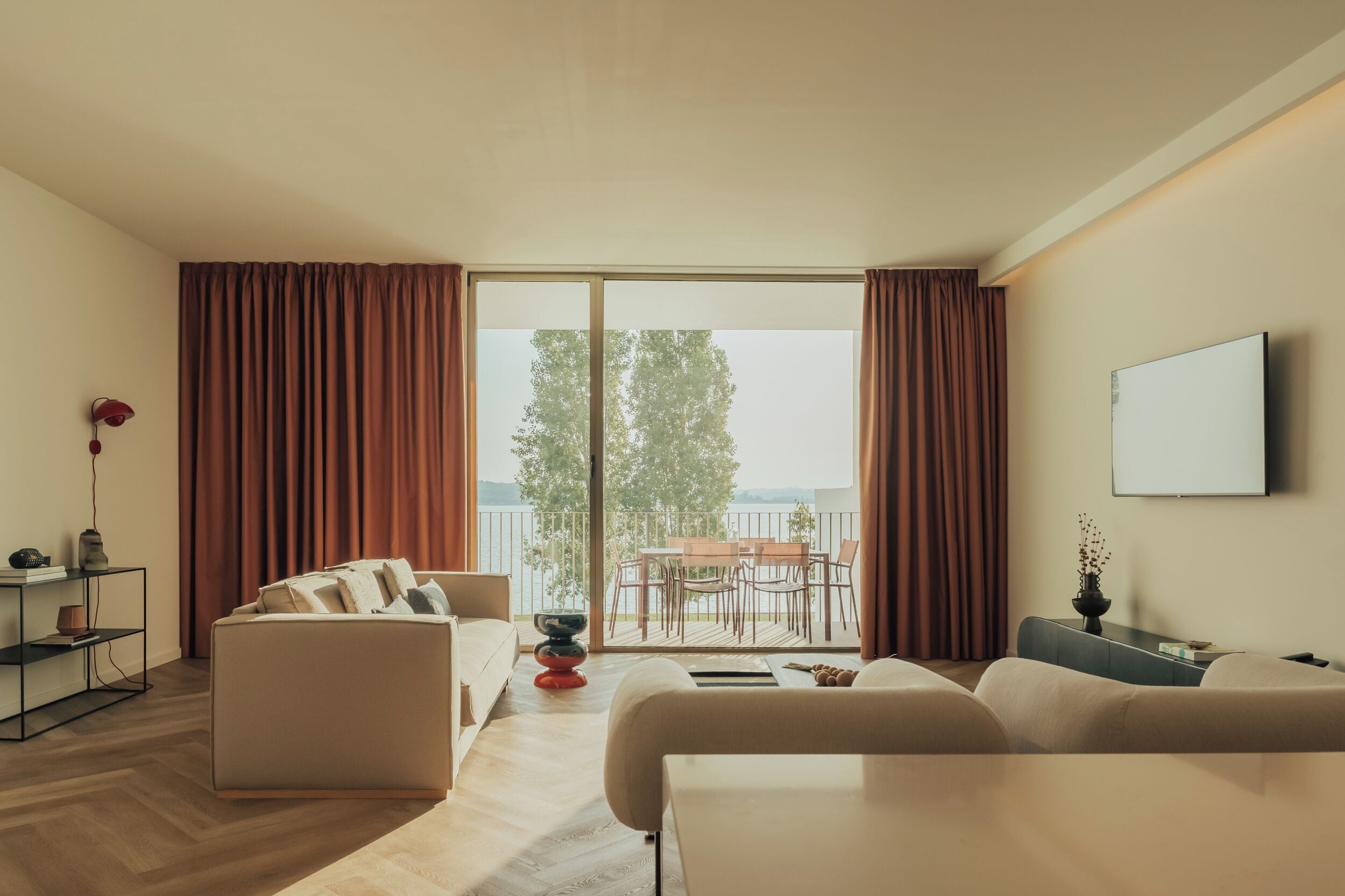 A modern living room with a sofa, a coffee table, and a dining area with chairs and a table. There is a large window with curtains, a flat-screen TV mounted on the wall, and a view of a balcony with outdoor furniture.