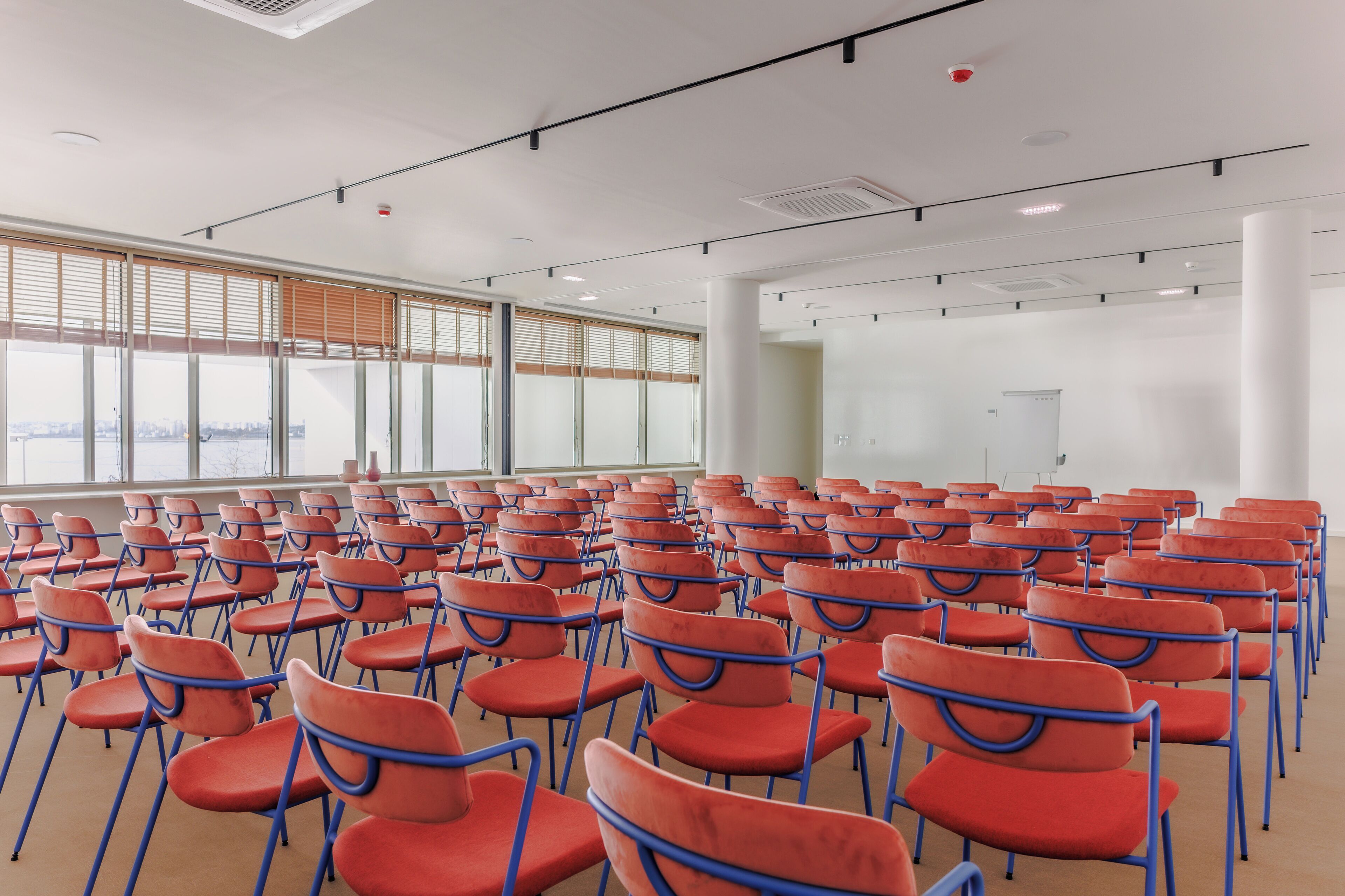 Un salón de conferencias con filas de sillas anaranjadas y un amplio ventanal con persianas.