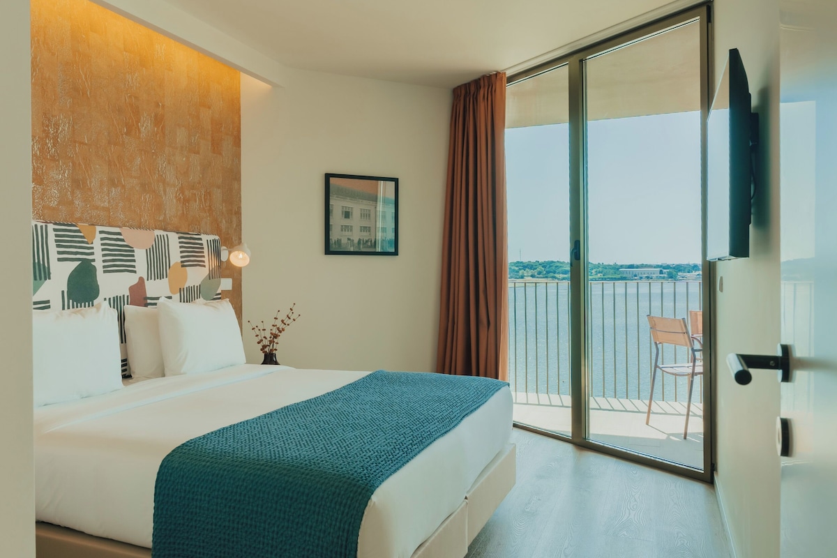 Habitación de hotel con cama, vista al mar, balcón con una silla y un cuadro en la pared.