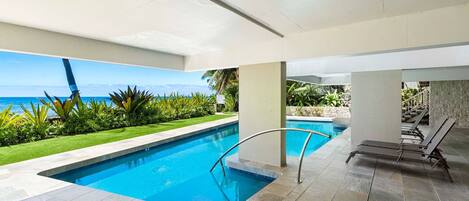 A swimming pool with a view of the ocean, surrounded by greenery and a tiled deck.