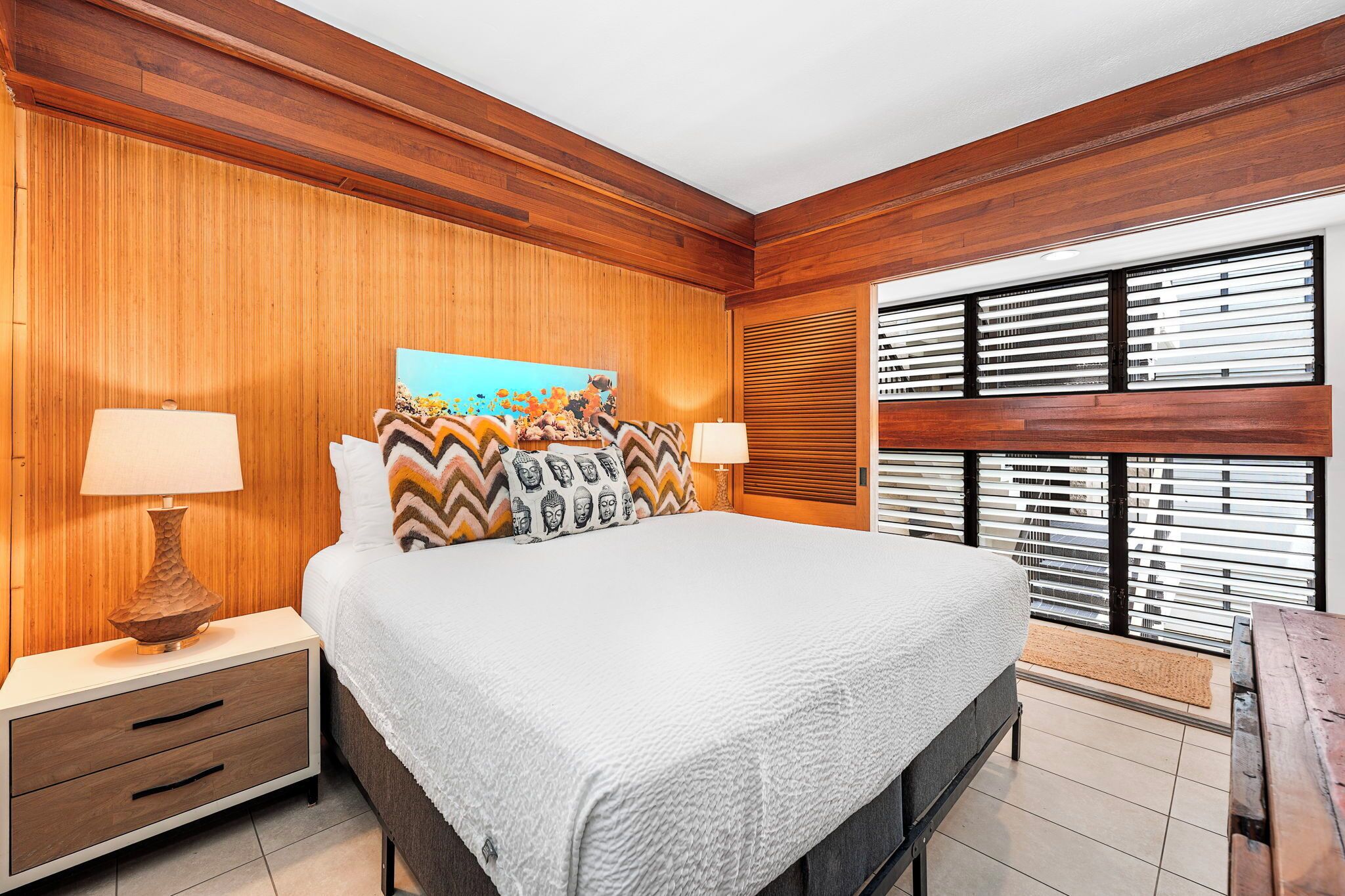 A bedroom with a wooden wall panel, a bed with patterned pillows, two bedside lamps, and a window with blinds.