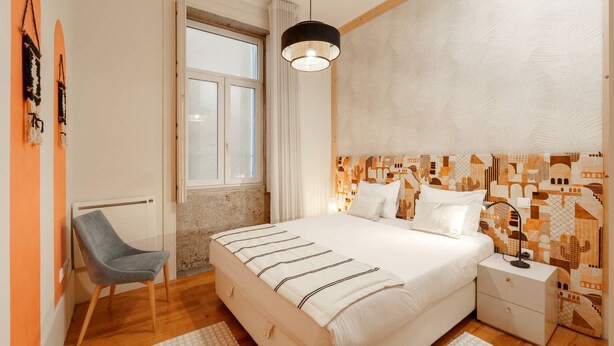 Un dormitorio con una cama, una mesita de noche, una silla, un ventanal y un papel tapiz estampado.