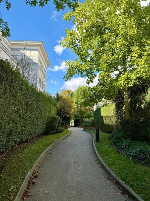 Uma trilha pavimentada, ladeada por vegetação densa e um edifício com fachada branca.