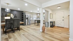 Una cocina moderna con electrodomésticos de acero inoxidable, un comedor con mesa y sillas de madera, y un pasillo que conduce a una puerta blanca.