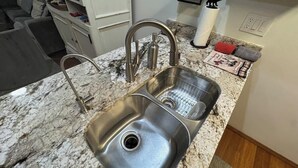 Una cocina con un lavamanos doble de acero inoxidable, encimera de granito y una pequeña repisa con utensilios de cocina.