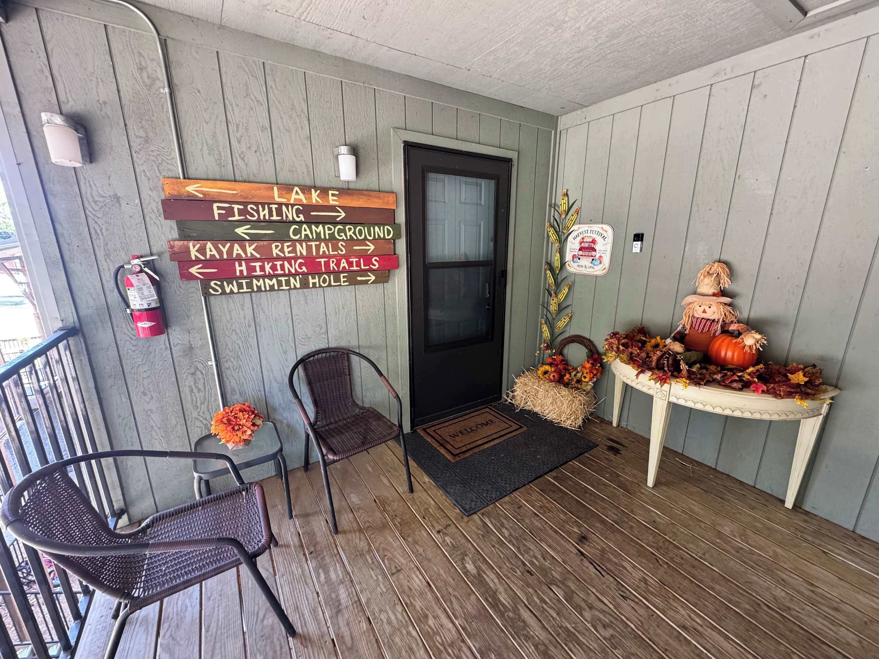 A wooden porch with a welcome mat, two wicker chairs, a table with autumn decorations, and directional signs for various activities.