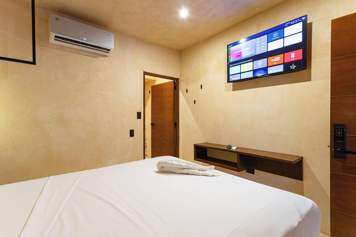 Habitación de hotel con cama, unidad de aire acondicionado, televisor integrado mostrando diversas aplicaciones, puerta de madera y estante empotrado.