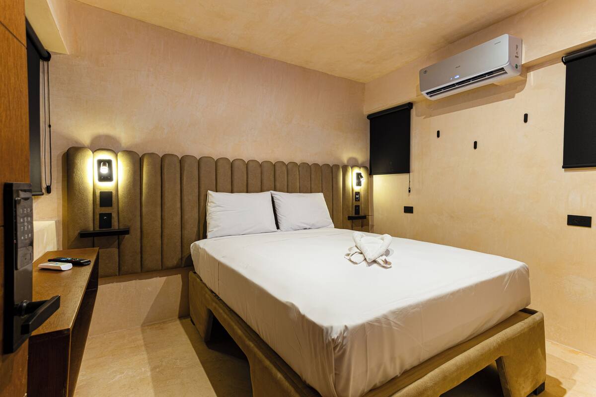 Habitación de hotel con cama, mesitas de noche, unidad de aire acondicionado y lámpara fijada a la pared.