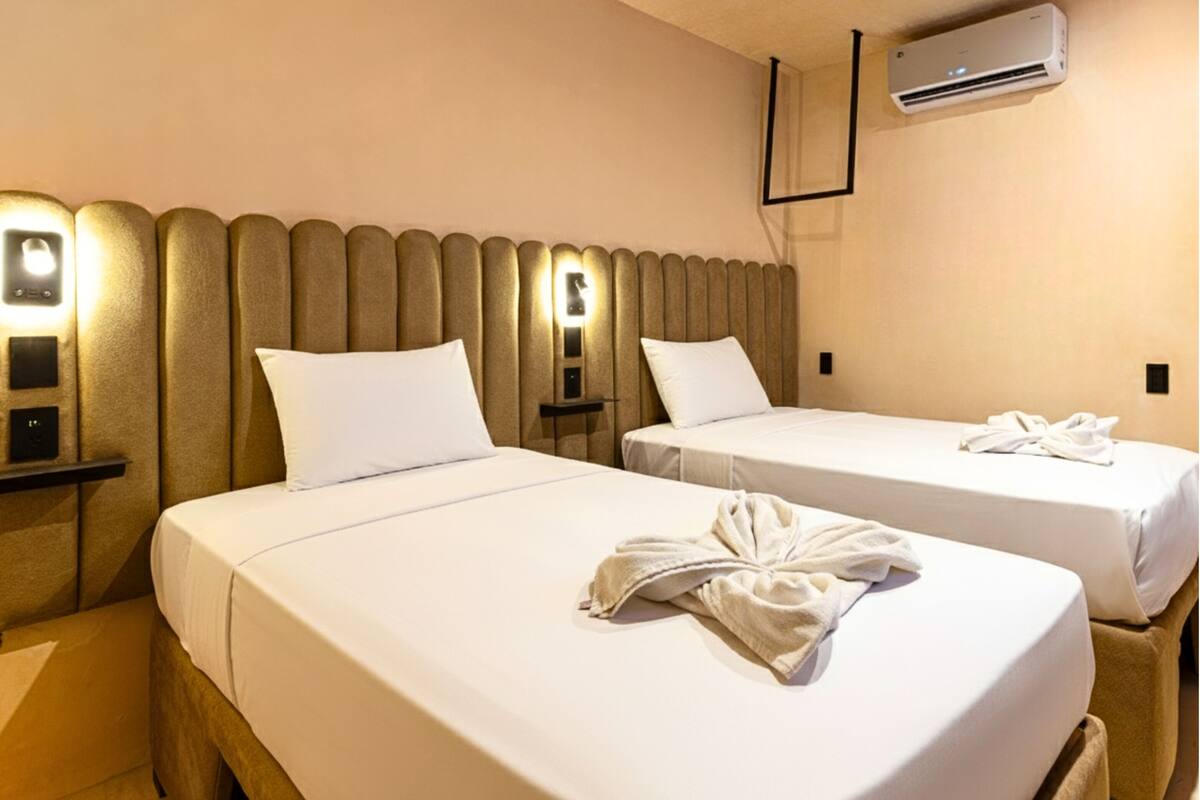 Habitación de hotel con dos camas, cada una con ropa y almohadas blancas, y una toalla sobre una cama. Se observa un aire acondicionado de pared y una mesita de noche con lámpara entre las camas.