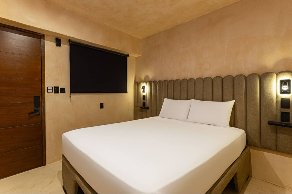 Una habitación de hotel con una cama grande, una puerta de madera, un televisor montado en la pared y un cabecero con iluminación integrada.