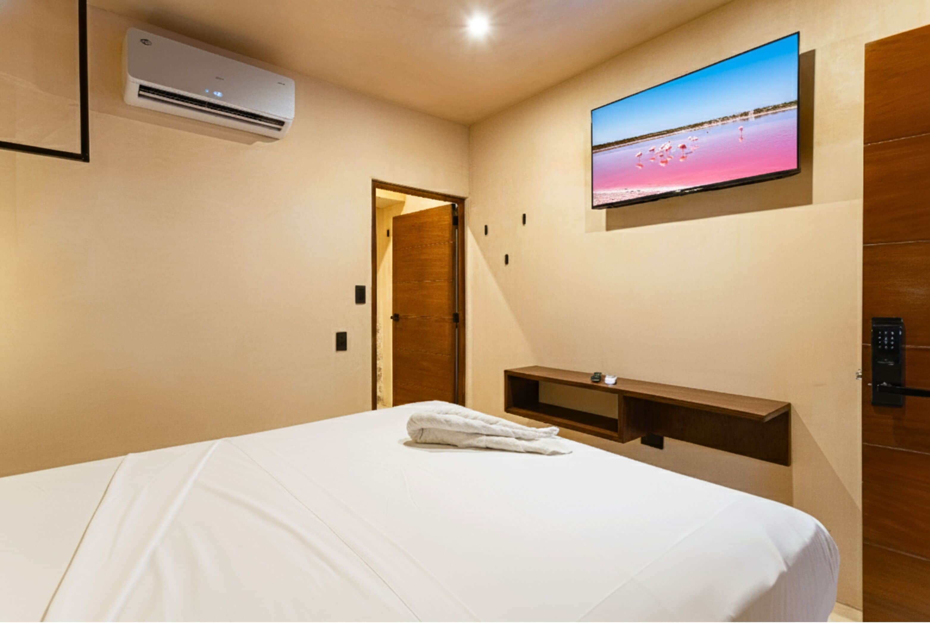 A hotel room with a bed, an air conditioner, a TV mounted on the wall, and a wooden door.