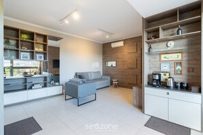 A modern living room with a grey sofa, a small side table, and a built-in kitchen area with a coffee machine and shelves. - Apt w/ Gourmet Balcony 300m From Beach Cms0202 (Florianópolis)