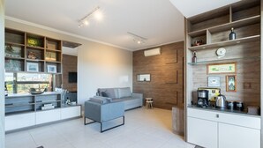 A modern living room with a grey sofa, a small side table, and a built-in kitchen area with a coffee machine and shelves.
