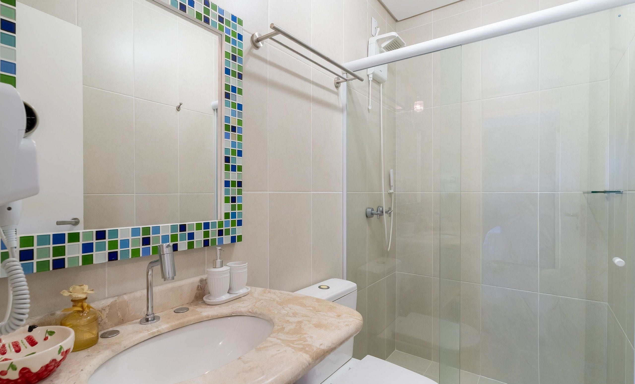 Casa de banho com cabine de duche em vidro, lavatório com bancada de mármore e espelho com moldura de mosaicos.