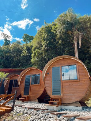 Une rangée de cabanes cylindriques en bois, dotées de fenêtres et de portes, est installée sur un sol de gravier, avec des arbres en arrière-plan.