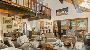 Un'ampia zona giorno con soffitto alto, una scala in legno, una cucina con elettrodomestici moderni e un'ampia porta a vetri scorrevoli che dà su un patio esterno.