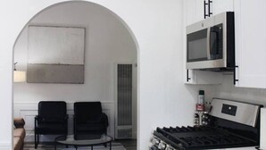Cozinha moderna com armários brancos, micro-ondas embutido e fogão a gás. Há uma área de estar com duas cadeiras e uma mesa, e um tapete estampado no chão.