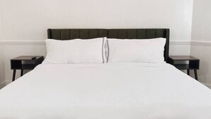 Cama com cabeceira escura e roupa de cama branca num quarto com chão de madeira.