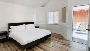 Quarto com piso de madeira, cama com roupa de cama branca, mesa de cabeceira, janela com persianas e porta que dá para uma área externa.