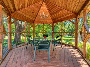 A wooden gazebo with a glass enclosure, a table, and chairs, surrounded by a wooden deck and trees.