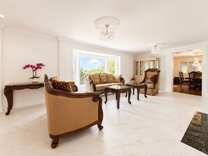 A spacious living room with a large window, a chandelier, and a dining area with wooden furniture.
