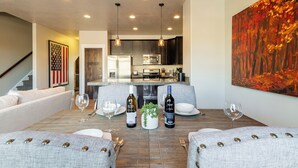 A dining area with a wooden table, two grey upholstered chairs, a chandelier, and a kitchen in the background.
