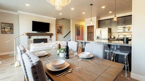A modern dining area with a wooden table set for four, a fireplace, a television, and a kitchen in the background.
