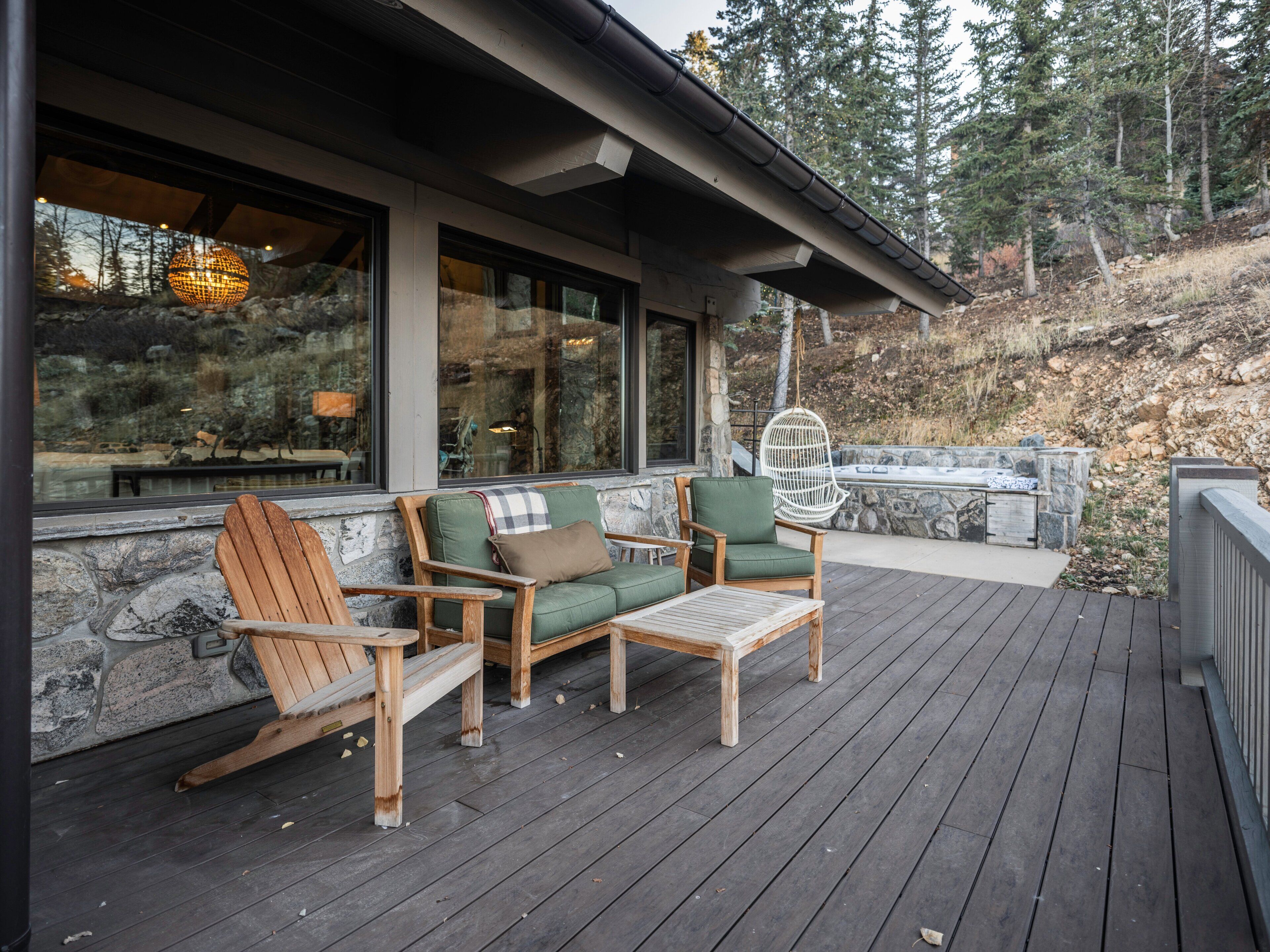 Una terraza de madera con mobiliario exterior, que incluye una mecedora y dos sofás, con vistas a un muro de piedra y una ladera boscosa.