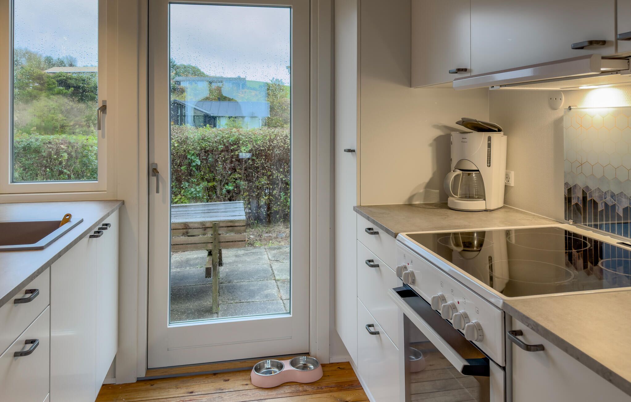 Une cuisine moderne avec des meubles blancs, une machine à café et une porte vitrée donnant sur une terrasse.