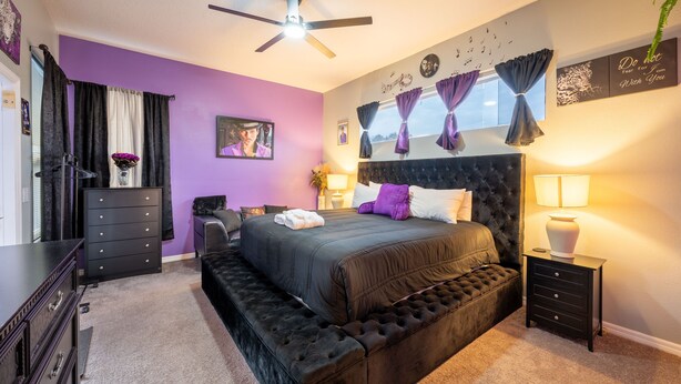 A bedroom with a large bed, a ceiling fan, a dresser, and a nightstand with a lamp.
